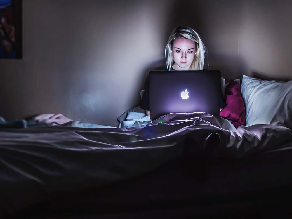 Frau sitzt im Dunkeln auf ihrem Bett mit Macbook auf dem Schoß